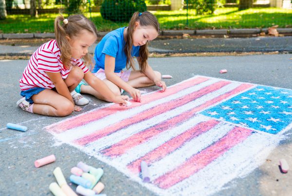 Two cute friends drawing American flag with colored chalks on the sidewalk near the house on sunny summer day.
