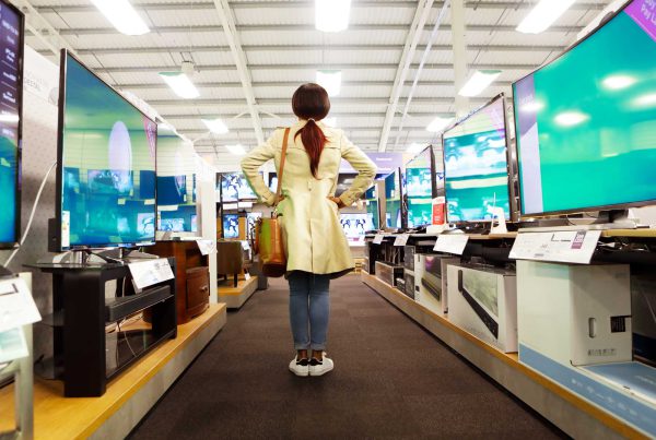 A woman in a light trench coat stands akimbo with her back to us and stares down the middle of a store's TV aisle.