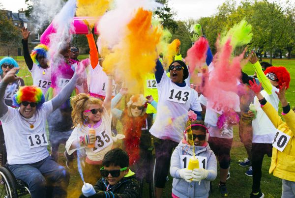 Kids, adults, a wheelchair user, and friends in race bibs and neon sunglasses celebrate with Holi powder at the end of a charity race.