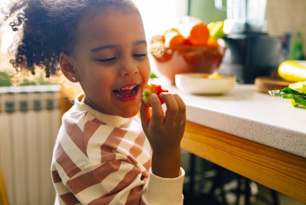 A little Black girl hold a strawberry up to her mouth and smiles after taking a bite.