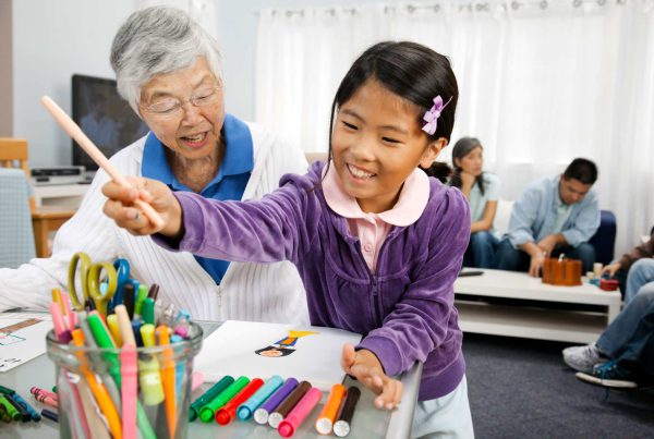 An Asian grandmother watches as her granddaughter uses markers to draw a picture.