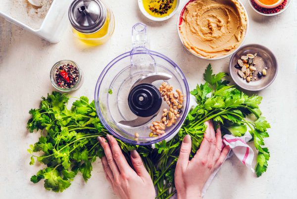 A woman's hands arrange herbs next to a food processor bowl filled with chickpeas. Olive oil and other ingredients sit nearby on the table.