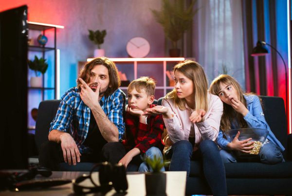 A family of four sits on the sofa and boredly looks at the TV.