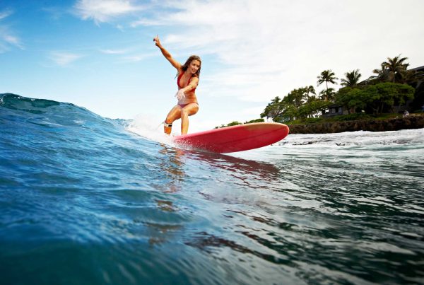 A woman rides an orange surfboard on a small wave with partly cloudy skies and palm trees in the background.