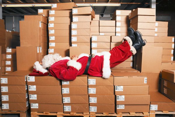 Santa Claus lays atop stacks of boxes waiting to be mailed. His feet are propped up. Looks like he's taking a nap.