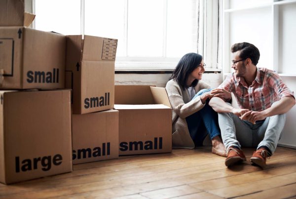 A couple sits on the floor on an empty apartment surrounded by moving boxes.