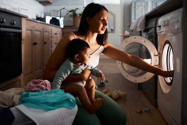 A mom with an infant on her hip pulls laundry from the dryer in a dark kitchen.
