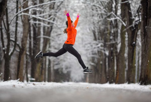 A woman in running clothes and a bright orange pullover leaps across a snowy walkway lined with snow-covered trees.