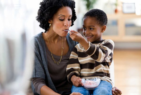 A child sits on his mom's lap as they share a bowl of ice cream.