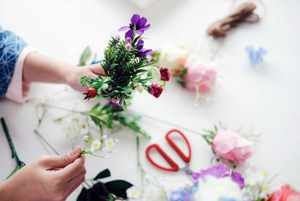 A close up of two hands, one holding a small bouquet, the other holding some ribbon, over a while table strewn with crafting supplies like scissors, twine, and individual faux floral stems.